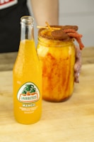 A bottle of mango-flavored Jarritos soda is positioned on a wooden surface next to a large jar filled with a vibrant mango drink. The jar features a spiced rim and is garnished with skewers of chili-covered candies. A person is holding the jar with their left hand.
