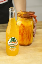 A bottle of mango-flavored Jarritos soda is positioned on a wooden surface next to a large jar filled with a vibrant mango drink. The jar features a spiced rim and is garnished with skewers of chili-covered candies. A person is holding the jar with their left hand.