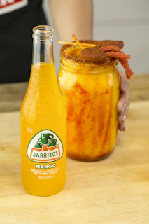 Bright glass jar filled with tangy mango achaar surrounded by fresh mangoes and spices.