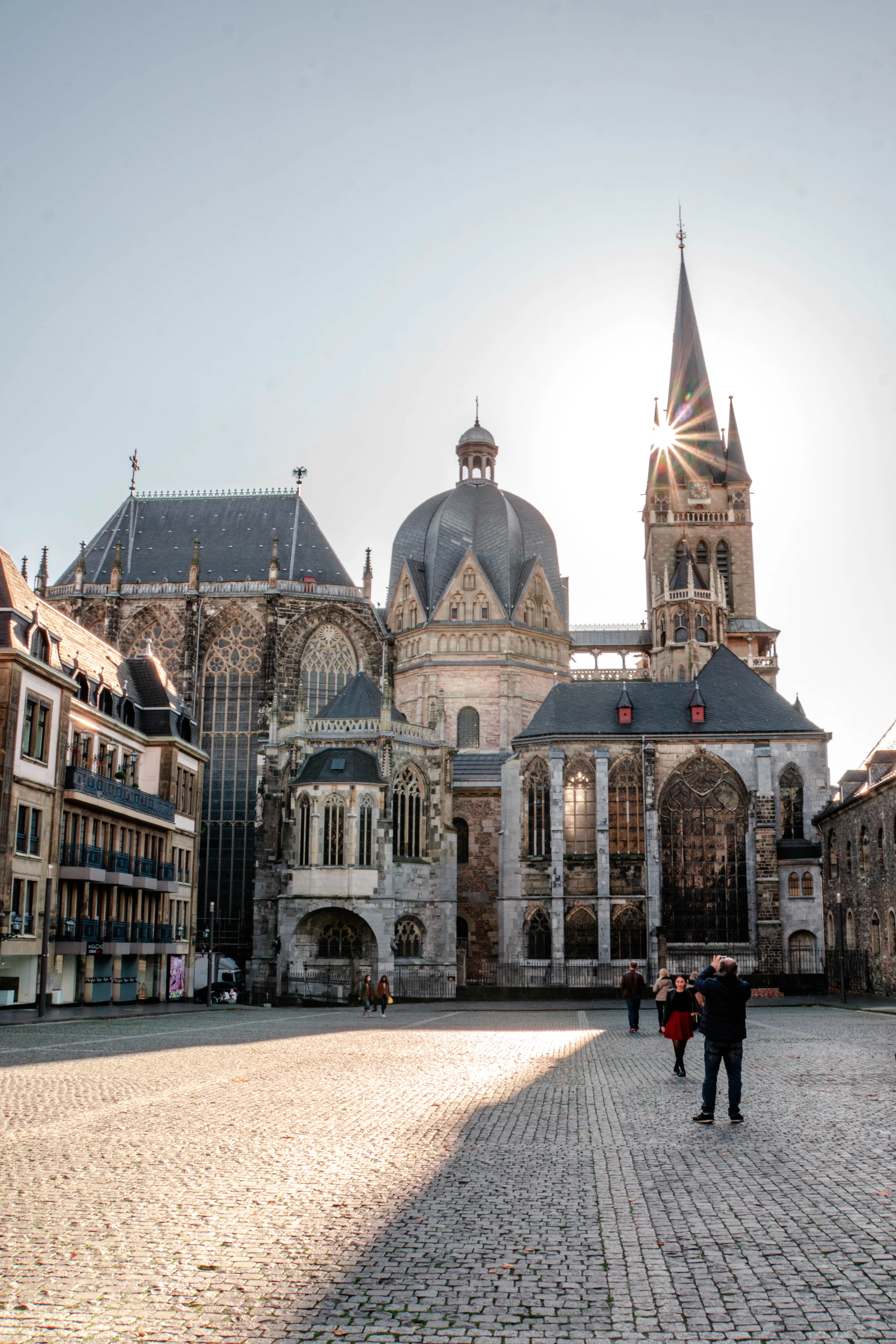 Aachen Cathedral seen across the cobbles of the Katschhof, its octagonal Carolingian dome and steep slate roofs in silhouette as the sun bursts from behind the spire of the west tower on a cold clear morning.