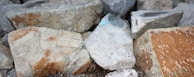 Several large, rough stones of varying sizes and colors are piled together. The stones have an uneven texture and display natural patterns and colors, including shades of gray, brown, and hints of blue.