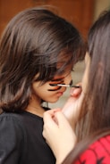 A young girl is having her face painted with black and orange stripes by another person. The focus is on the artistic application of the paint, capturing a close-up view of the hands and brush. The girl's hair is long and brown, and she is wearing a black top.