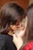 A young girl is having her face painted with black and orange stripes by another person. The focus is on the artistic application of the paint, capturing a close-up view of the hands and brush. The girl's hair is long and brown, and she is wearing a black top.