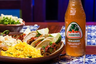 A wooden table features vibrant, colorful Mexican cuisine, including tacos with lime wedges, cooked rice with herbs, and coleslaw. Beside the plate, there is a bottle of Jarritos Tamarind soda, characterized by its amber color and distinctive label.