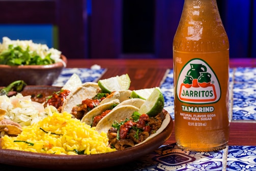 A wooden table features vibrant, colorful Mexican cuisine, including tacos with lime wedges, cooked rice with herbs, and coleslaw. Beside the plate, there is a bottle of Jarritos Tamarind soda, characterized by its amber color and distinctive label.