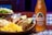 A wooden table features vibrant, colorful Mexican cuisine, including tacos with lime wedges, cooked rice with herbs, and coleslaw. Beside the plate, there is a bottle of Jarritos Tamarind soda, characterized by its amber color and distinctive label.