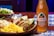 A wooden table features vibrant, colorful Mexican cuisine, including tacos with lime wedges, cooked rice with herbs, and coleslaw. Beside the plate, there is a bottle of Jarritos Tamarind soda, characterized by its amber color and distinctive label.
