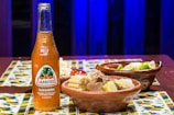 A bottle of Jarritos tamarind soda is placed on a wooden table with decorative tiles. Beside it, there are clay bowls filled with lime wedges and a dish containing corn, meat, and vegetables.