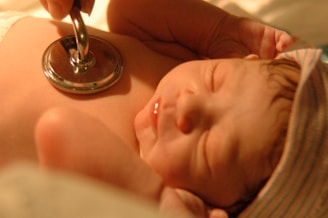 A newborn baby is lying peacefully while a stethoscope is placed on its chest, symbolizing a medical check-up. The baby is wearing a striped cap and has a serene expression, illuminated by warm lighting.