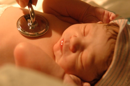 A newborn baby is lying peacefully while a stethoscope is placed on its chest, symbolizing a medical check-up. The baby is wearing a striped cap and has a serene expression, illuminated by warm lighting.