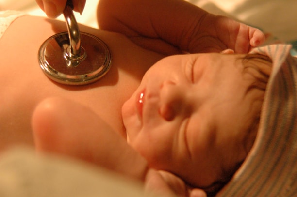 A newborn baby is lying peacefully while a stethoscope is placed on its chest, symbolizing a medical check-up. The baby is wearing a striped cap and has a serene expression, illuminated by warm lighting.