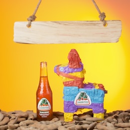 A vibrant piñata in colorful paper shades of red, blue, yellow, and purple sits to the right of an orange Jarritos soda bottle on a background filled with tamarind pods. Above them, a simple wooden sign hangs from two ropes. The background is a bright gradient of yellow and orange, adding a festive atmosphere.
