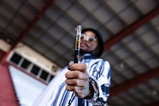 A person in a striped shirt and sunglasses holds a vaping device prominently in the foreground. The background consists of a roof with metal beams, giving an industrial vibe.