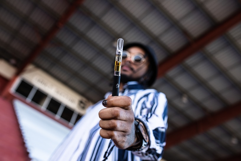 A person in a striped shirt and sunglasses holds a vaping device prominently in the foreground. The background consists of a roof with metal beams, giving an industrial vibe.