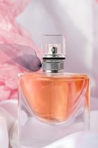 A clear glass perfume bottle with an orange-colored liquid inside is topped with a sleek silver cap and decorated with a delicate dark ribbon. The background is adorned with soft pink and white drapery, enhancing the luxurious and elegant feel.