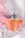 A clear glass perfume bottle with an orange-colored liquid inside is topped with a sleek silver cap and decorated with a delicate dark ribbon. The background is adorned with soft pink and white drapery, enhancing the luxurious and elegant feel.