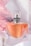 A clear glass perfume bottle with an orange-colored liquid inside is topped with a sleek silver cap and decorated with a delicate dark ribbon. The background is adorned with soft pink and white drapery, enhancing the luxurious and elegant feel.