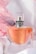 A clear glass perfume bottle with an orange-colored liquid inside is topped with a sleek silver cap and decorated with a delicate dark ribbon. The background is adorned with soft pink and white drapery, enhancing the luxurious and elegant feel.