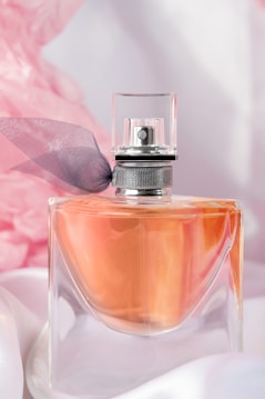 A clear glass perfume bottle with an orange-colored liquid inside is topped with a sleek silver cap and decorated with a delicate dark ribbon. The background is adorned with soft pink and white drapery, enhancing the luxurious and elegant feel.
