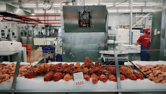 A seafood market display featuring freshly cooked crayfish laid out on ice, with various other seafood items surrounding them. Behind the counter, a worker wearing a red shirt and cap is attending to tasks, and industrial kitchen equipment is visible in the background.