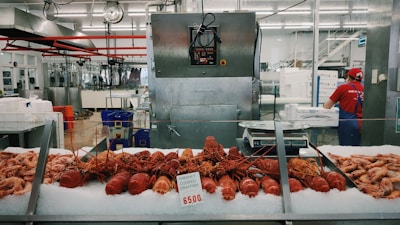 A seafood market display featuring freshly cooked crayfish laid out on ice, with various other seafood items surrounding them. Behind the counter, a worker wearing a red shirt and cap is attending to tasks, and industrial kitchen equipment is visible in the background.