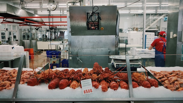 A seafood market display featuring freshly cooked crayfish laid out on ice, with various other seafood items surrounding them. Behind the counter, a worker wearing a red shirt and cap is attending to tasks, and industrial kitchen equipment is visible in the background.