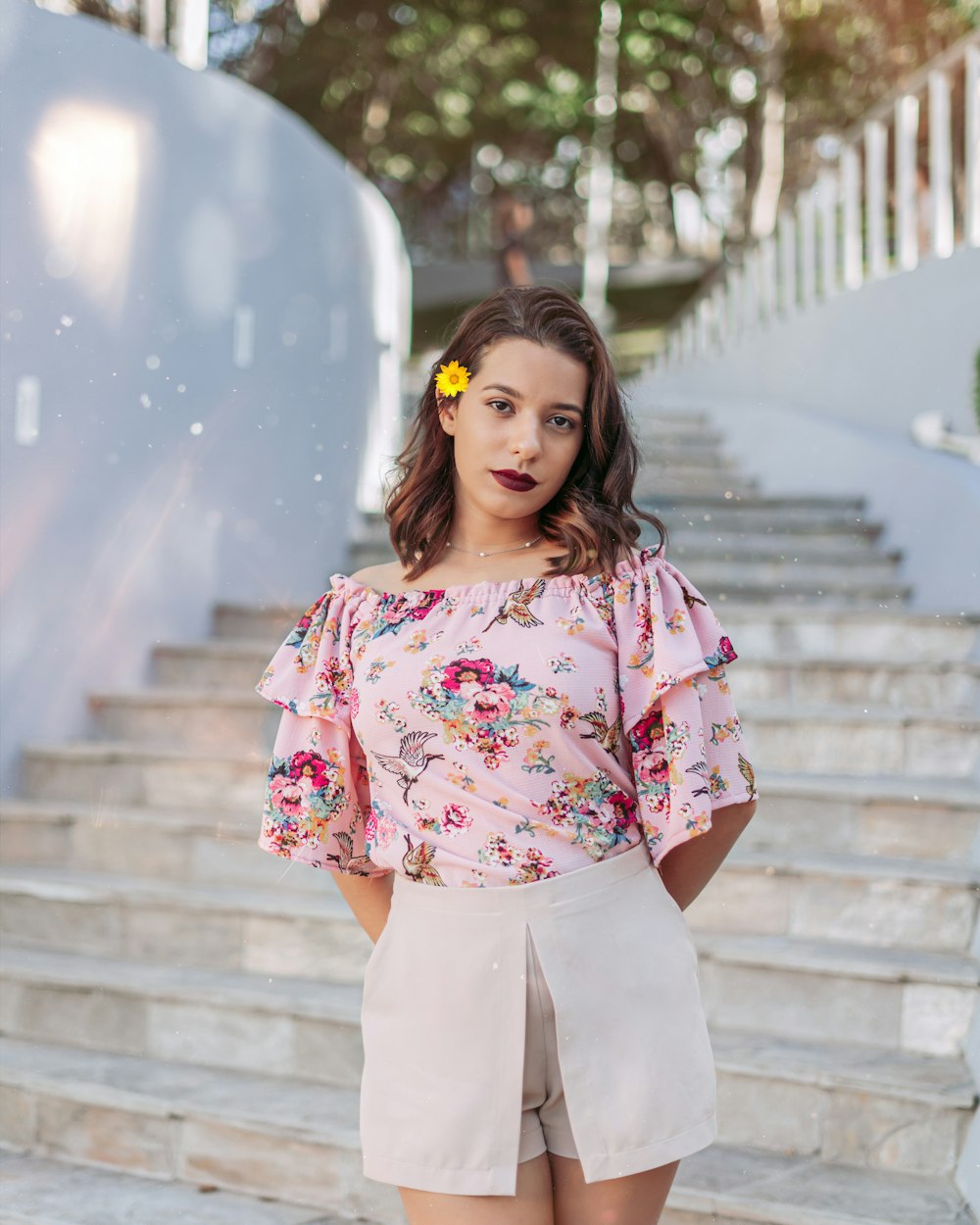 Femme en chemise à fleurs rose et blanche et pantalon blanc debout sur des escaliers en béton