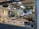 An artist's studio filled with various artworks, including framed paintings and sketches displayed on the walls and a countertop. Architectural models and design pieces are spread across the workspace. A large green plant adds a natural element, and studio lighting creates a focused atmosphere.