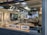 An artist's studio filled with various artworks, including framed paintings and sketches displayed on the walls and a countertop. Architectural models and design pieces are spread across the workspace. A large green plant adds a natural element, and studio lighting creates a focused atmosphere.