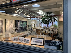 An artist's studio filled with various artworks, including framed paintings and sketches displayed on the walls and a countertop. Architectural models and design pieces are spread across the workspace. A large green plant adds a natural element, and studio lighting creates a focused atmosphere.