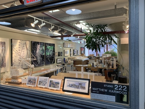 An artist's studio filled with various artworks, including framed paintings and sketches displayed on the walls and a countertop. Architectural models and design pieces are spread across the workspace. A large green plant adds a natural element, and studio lighting creates a focused atmosphere.