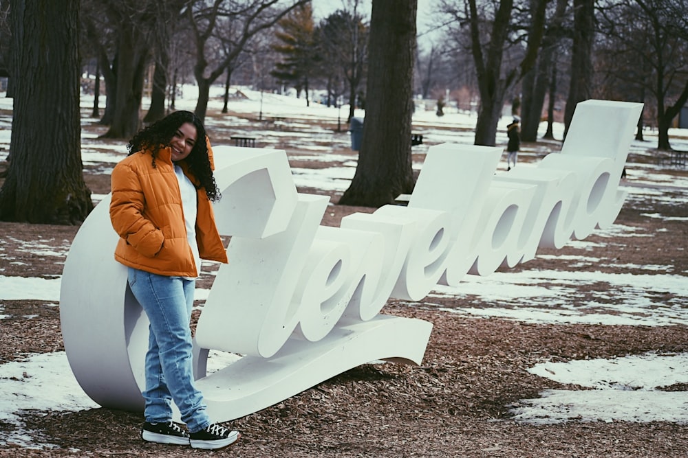 woman in orange jacket and blue denim jeans standing near white wooden bench during daytime