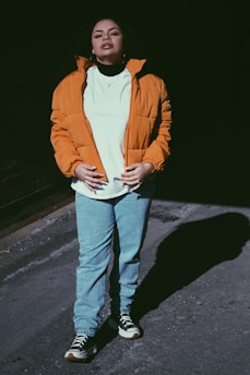 A person stands confidently outdoors in a casual outfit, wearing a bright orange jacket over a white T-shirt paired with blue jeans and sneakers. Their expression suggests confidence and calmness. Shadows cast by sunlight create a dramatic effect on the ground.