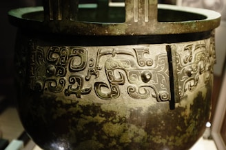 An intricately designed bronze vessel featuring elaborate swirling patterns and motifs. The vessel appears ancient, with the designs showing a high level of craftsmanship, typical of traditional art. The surface has a patina, indicating its age and historical significance.