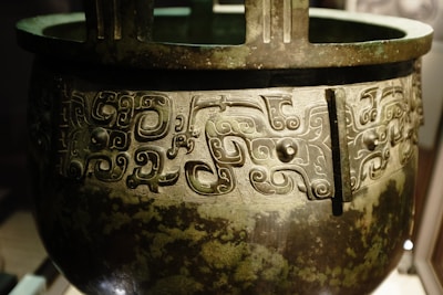 An intricately designed bronze vessel featuring elaborate swirling patterns and motifs. The vessel appears ancient, with the designs showing a high level of craftsmanship, typical of traditional art. The surface has a patina, indicating its age and historical significance.