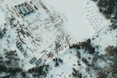 An aerial view of a snowy landscape with several large vehicles, likely trucks or trailers, organized in a patterned manner. The ground is partially covered with snow, revealing patches of the underlying earth. Trees are scattered around the perimeter, adding a touch of greenery to the predominantly white scene.