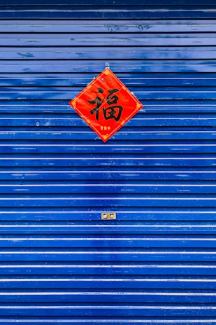 A blue metal shutter with horizontal lines features a red diamond-shaped piece of paper with Chinese characters in the center. The surface of the shutter has a glossy finish, with some reflections visible.