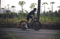 A person wearing a cycling helmet and dark athletic wear is riding a bicycle on a paved road. The background features lush greenery with thin tall trees and mountains in the distance during what appears to be early morning or late afternoon.