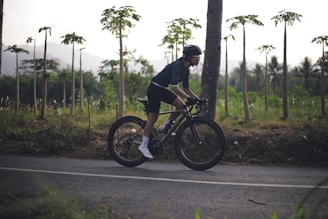 A person wearing a cycling helmet and dark athletic wear is riding a bicycle on a paved road. The background features lush greenery with thin tall trees and mountains in the distance during what appears to be early morning or late afternoon.