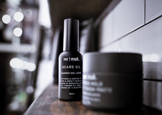 A sleek bottle of beard oil positioned prominently on a wooden shelf, with another container blurred in the background. The setting appears modern and minimalistic, featuring white tiles on the wall.