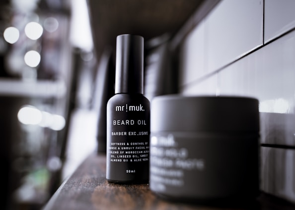 A sleek bottle of beard oil positioned prominently on a wooden shelf, with another container blurred in the background. The setting appears modern and minimalistic, featuring white tiles on the wall.