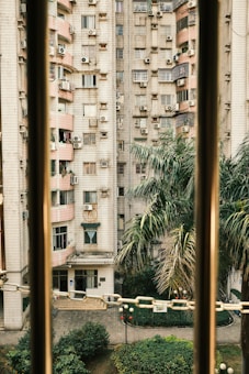 A view of a residential building with numerous air conditioning units attached to the exterior walls. The building features a pattern of square tiles and several balconies with various items placed outside. Palm trees and other greenery are visible in the foreground, indicating an urban environment with landscaped areas. Part of the image is framed by vertical bars and a chain.