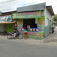A small juice stall with colorful signage displaying various fruit juice options and prices. A man on a motorcycle and a woman are present in front of the stall. The stall is part of a larger, simple building with a corrugated metal roof. The scene includes other shops with closed green shutters.