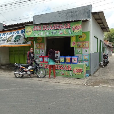 A small juice stall with colorful signage displaying various fruit juice options and prices. A man on a motorcycle and a woman are present in front of the stall. The stall is part of a larger, simple building with a corrugated metal roof. The scene includes other shops with closed green shutters.