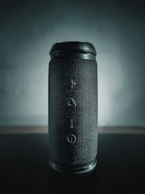 A cylindrical portable speaker with a textured grille, vertical buttons for volume, play, and power arranged centrally. The lighting creates a soft gradient background with a focus on the speaker.