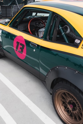 A vintage-style car with a distinctive yellow and green paint job, featuring a prominent pink circle with the number 13 on the door. The car has a retro design with a visible roll cage inside, a red racing steering wheel, and golden alloy wheels. The setting suggests a motorsport or racing context.