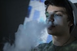 A person exhales a large cloud of smoke in a dimly lit environment. The background features a tiled wall with bluish-white lighting. The person is wearing a beanie and looks calm and pensive.