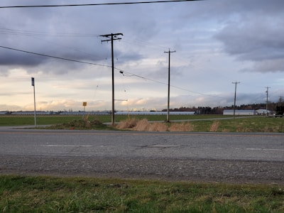 A rural landscape with a road, fields, and a series of power lines stretching across the scene. The foreground features a grassy verge next to the road. In the background, there are industrial buildings and distant trees under a sky filled with clouds.