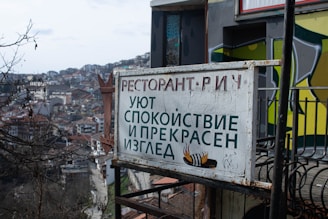A weathered sign featuring text in Cyrillic script sits in the foreground, attached to a metal frame. It is slightly rusted and features a graphic of steaming food. The background shows a densely packed urban landscape with numerous buildings on a hillside, and a cloudy sky above.