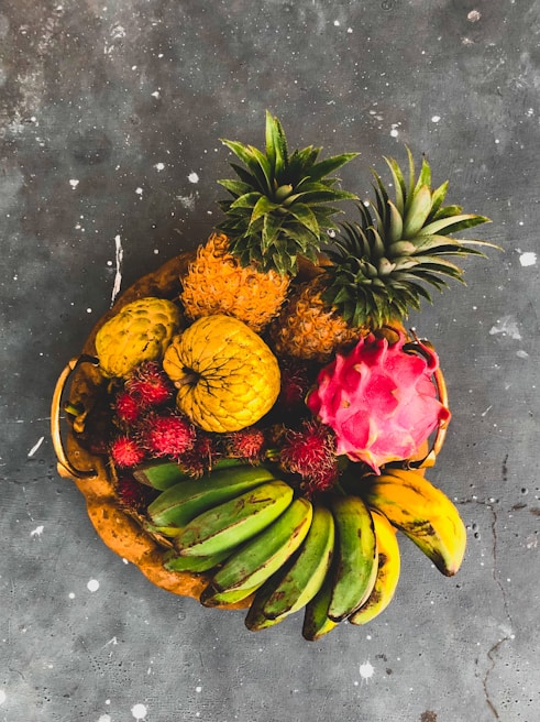 A vibrant arrangement of tropical fruits including bananas, pineapples, rambutan, dragon fruit, and a soursop, displayed in a wicker basket on a textured dark surface.